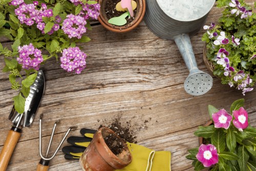 A close-up image of a wooden outdoor table used for gardening, featuring several potted flowering plants with pink and purple blooms, a metal watering can with a long spout, small gardening tools including a hand rake, a trowel, and a pair of gardening gloves. The scene also includes a small empty flower pot, soil, and decorative wooden plant markers. The background suggests a typical outdoor garden setting, indicating an outdoor space in Maida Vale suitable for gardening services such as planting, potting, and general outdoor maintenance. The lighting appears natural, highlighting the vibrant colours of the flowers and the rustic texture of the wooden surface, supporting the context of a professional gardening environment.