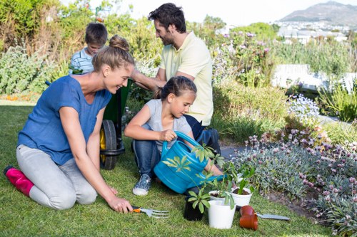 A family gardening in a well-maintained backyard within a British residential garden, featuring a lush green lawn, various flowering plants, and neatly bordered flower beds. The foreground shows a woman in a blue top and grey trousers kneeling on the grass, planting or tending to a young shrub with a small garden fork nearby. A small girl with a backpack is actively watering plants with a watering can, while other gardening tools and pots are arranged around them. In the background, a man with dark hair, wearing a cream shirt, is assisting two children with their gardening activities amidst a backdrop of shrubs, flowering bushes, and trees. The outdoor space appears well-organized, with defined lawn areas, flower beds, and pathways, under natural light suggesting a bright, partly cloudy day. This scene aligns with professional gardening and landscaping services offered by Gardening Services Maida Vale, capturing the engaging and natural elements of a typical garden maintained for aesthetic and outdoor enjoyment in the local London area.